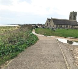a tarmac footpath with greenery and the sea on the left and leading to All Saints' and St Margaret's Church, Pakefield, on the right