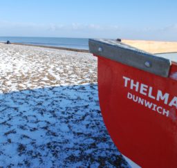 Dunwich Beach in winter - snow covers the shingle with fishermen's tents spread out along the beach. The stern of a bright red rowing bat called 