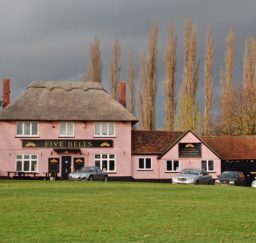 view of the Five Bells pub at Cavendish - a Suffolk pink thatched building with a long black barn attached, a green in the foreground, and tall trees behind, with a dramatic grey lowering sky