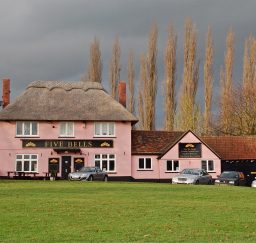 view of the Five Bells pub at Cavendish - a Suffolk pink thatched building with a long black barn attached, a green in the foreground, and tall trees behind, with a dramatic grey lowering sky
