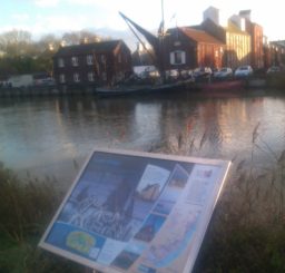 a view of Snape Maltings and a barge from the Sailors' Path, with an information board in the foreground