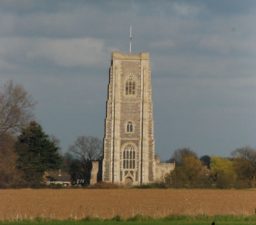 The grey flint square tower of Lavenham Church, with fields in the foreground and a cloudy sky in the background