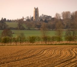 a sunset view across ploughed brown fields, a sweep of green field with lots of trees, and the square flint tower of Stoke by Nayland Church in the middle of the background