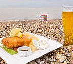 a delicious looking portion of fish, chips and mushy peas with a pint of beer on Aldeburgh Beach, with a blue and red striped windbreak and the sea in the background