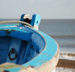 in focus taking up most of the picture is the prow of a tatty blue painted rowing boat, with the shingle beach and sea at Dunwich visible in the background